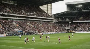 A wide view of a large professional European football stadium during a daytime match with a packed crowd and players
