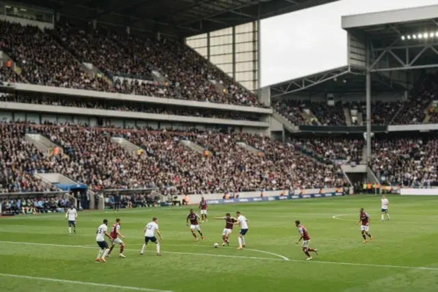 A wide view of a large professional European football stadium during a daytime match with a packed crowd and players